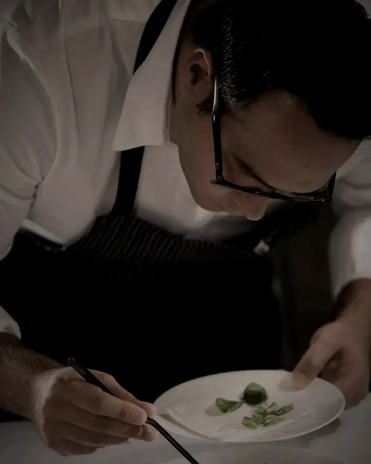 The chef bent over a plate, placing a leaf with tweezers under low light